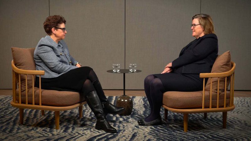 Two women sit facing each other on armchairs with cushions, separated by a small round table with two glasses of water, in a neutral-toned room.
