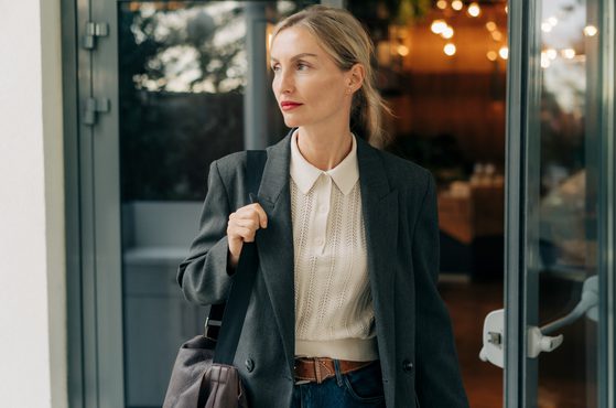 A woman in a dark grey blazer, white knit top, and jeans holding a black shoulder bag, standing near a glass door with a thoughtful expression.
