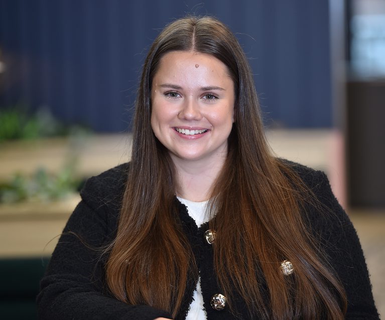 A smiling woman with long brown hair, wearing a black textured jacket with metal buttons, sitting indoors with a blurred background.