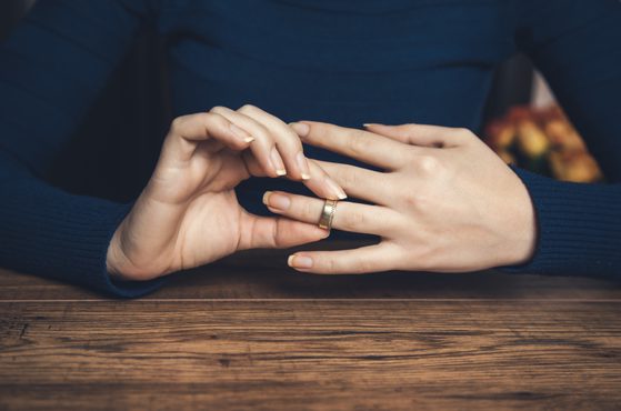 Hands resting on a wooden surface, with one hand touching a wedding ring on the other finger, wearing a dark blue long-sleeve shirt.