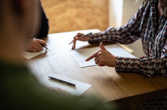 A person in a patterned long-sleeve shirt gesturing while sitting at a wooden table with papers and pens, engaged in a discussion with others.