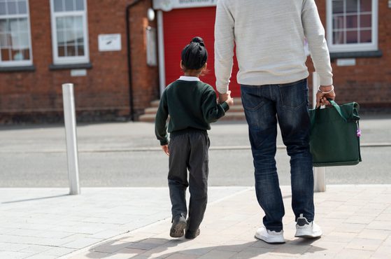 A man and a child walk hand in hand along a sidewalk outdoors, with the man holding a green bag in his other hand, in front of a brick building with red door.