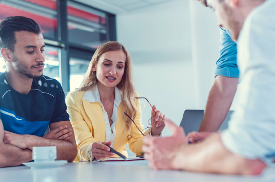 Four people are gathered around a table in a bright room, engaging in a discussion, with one woman holding glasses and a pen, and others listening attentively.