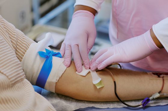 A healthcare professional in pink scrubs and pink gloves is inserting a needle into a patient's arm, which is secured with tape and a blue ribbon.