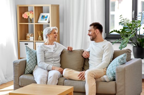 An elderly woman and a young man sit on a beige sofa, smiling and talking in a bright living room with a wooden bookshelf, potted plant, and large window.