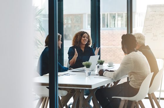 A woman with curly hair is speaking to a group of three colleagues in a modern office meeting room, with a whiteboard and large windows in the background.