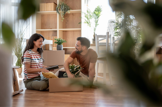 A young couple sits on the floor in a bright, plant-filled room, smiling at each other while unpacking a cardboard box filled with houseplants.