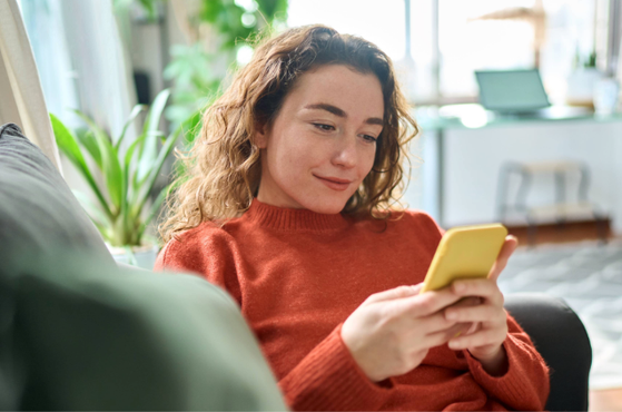 A woman with curly hair wearing an orange sweater sitting on a sofa and looking at her yellow smartphone in a bright living room.