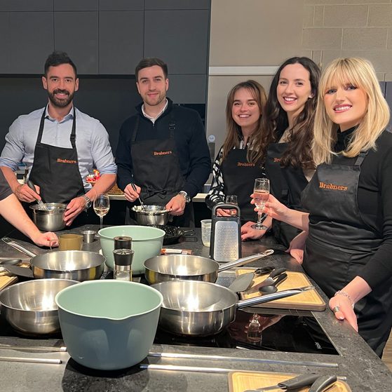 Six people in Brabners aprons stand around a modern kitchen island with pots, pans, cutting boards and utensils ready for cooking.