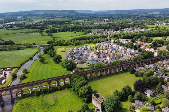 Aerial view of a small town with a viaduct running across green fields and a river, surrounded by lush trees, houses, and open countryside under a partly cloudy sky.
