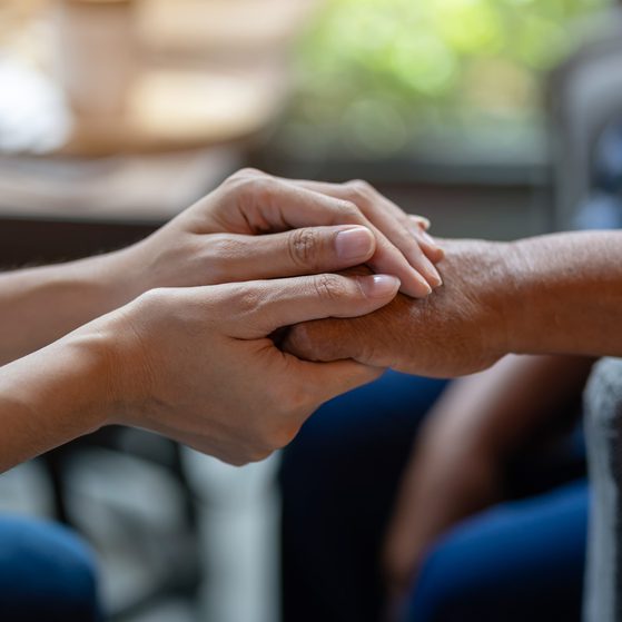 Two people holding hands, one gently supporting the other's wrist with a caring gesture. Indoor setting with natural light in the background.