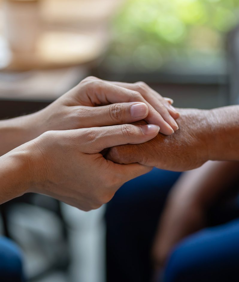 Two people holding hands, one gently supporting the other's wrist with a caring gesture. Indoor setting with natural light in the background.