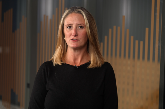 A woman with shoulder-length blonde hair wearing a black top, standing indoors with a modern, wooden office background featuring vertical slats.