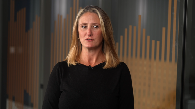 A woman with shoulder-length blonde hair wearing a black top, standing indoors with a modern, wooden office background featuring vertical slats.