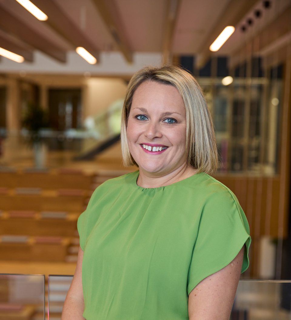 Smiling person with short blonde hair wearing a lime-green dress stands by a glass railing in a bright, open-plan, modern office interior.