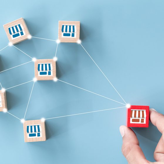 A hand placing a red wooden block with a shop icon onto a network of seven similar blocks with shop icons, connected by white lines on a light blue background.