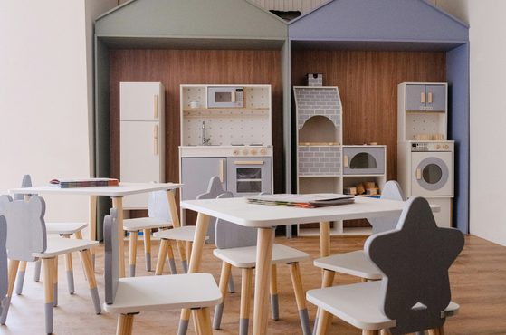 Bright playroom with two pastel toy kitchens set into alcoves, plus a fridge, oven and brick oven. White tables and grey-blue chairs on a wooden floor.