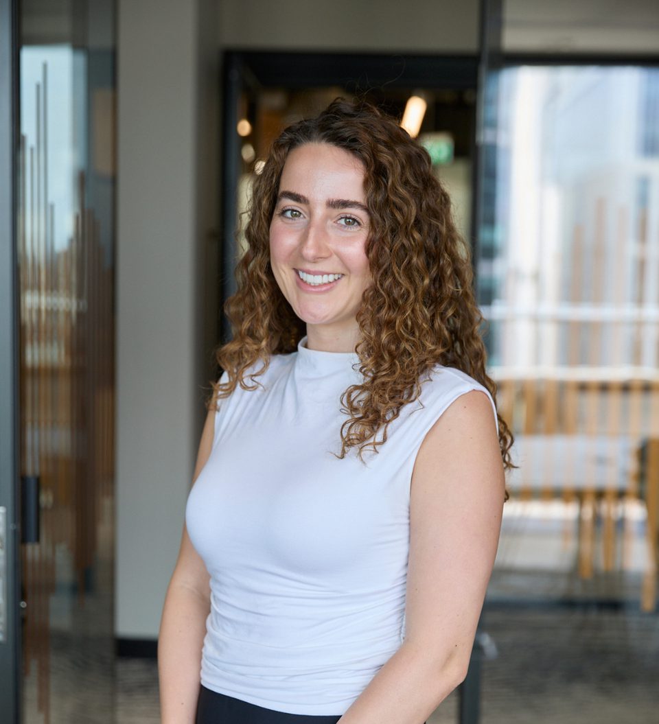 Person with long curly brown hair wearing a white sleeveless top, smiling at the camera, standing in a modern glass-walled office lobby interior.
