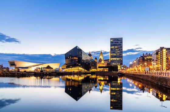 City skyline at dusk with modern black and glass buildings reflected in calm water, illuminated by streetlights and a partly cloudy sky.