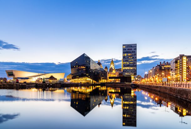 City skyline at dusk with modern black and glass buildings reflected in calm water, illuminated by streetlights and a partly cloudy sky.
