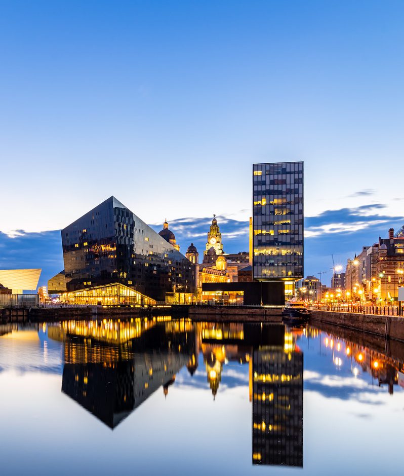 City skyline at dusk with modern black and glass buildings reflected in calm water, illuminated by streetlights and a partly cloudy sky.