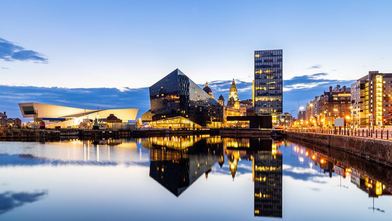 City skyline at dusk with modern black and glass buildings reflected in calm water, illuminated by streetlights and a partly cloudy sky.