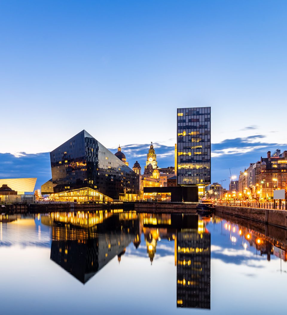 City skyline at dusk with modern black and glass buildings reflected in calm water, illuminated by streetlights and a partly cloudy sky.
