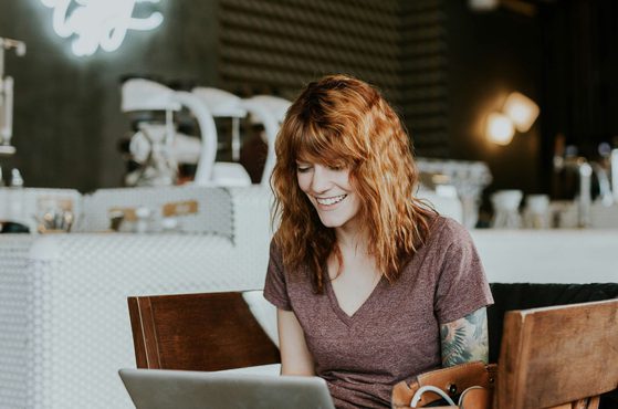 A woman with curly red hair, wearing a maroon t-shirt with a tattoo on her right arm, smiling while looking at a laptop in a cafe.