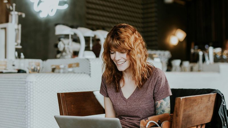 A woman with curly red hair, wearing a maroon t-shirt with a tattoo on her right arm, smiling while looking at a laptop in a cafe.