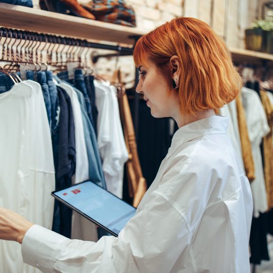 A woman with short red hair, wearing a white shirt, is shopping for clothes in a store, examining a white garment on a rack while holding a tablet.