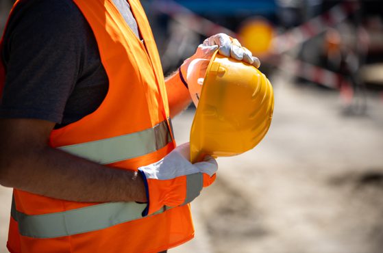 A person wearing an orange safety vest, grey shirt, and white gloves holding a yellow safety helmet at an outdoor construction site.