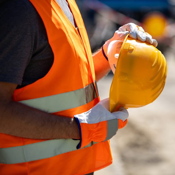 A person wearing an orange safety vest, grey shirt, and white gloves holding a yellow safety helmet at an outdoor construction site.
