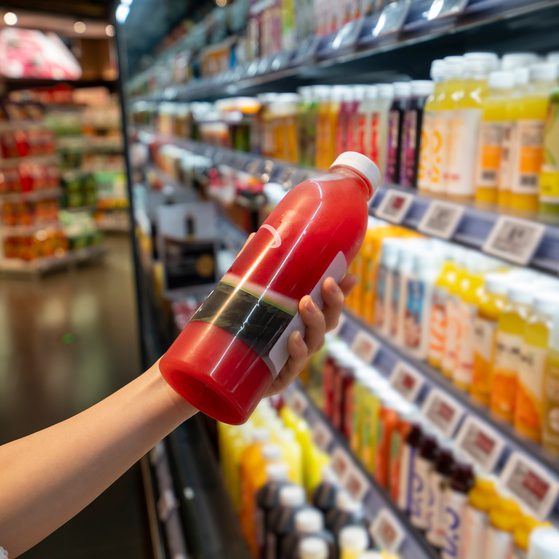 A person holds a red bottle with a white cap, browsing the shelves of a store filled with various bottles of drinks or supplements in the background.