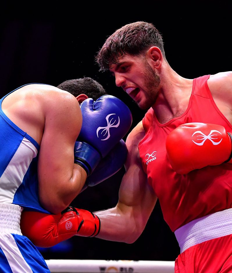 Two male boxers are engaged in a match; one in blue and the other in red. They are in close contact, with the boxer in blue covering his face while the boxer in red is throwing a punch.