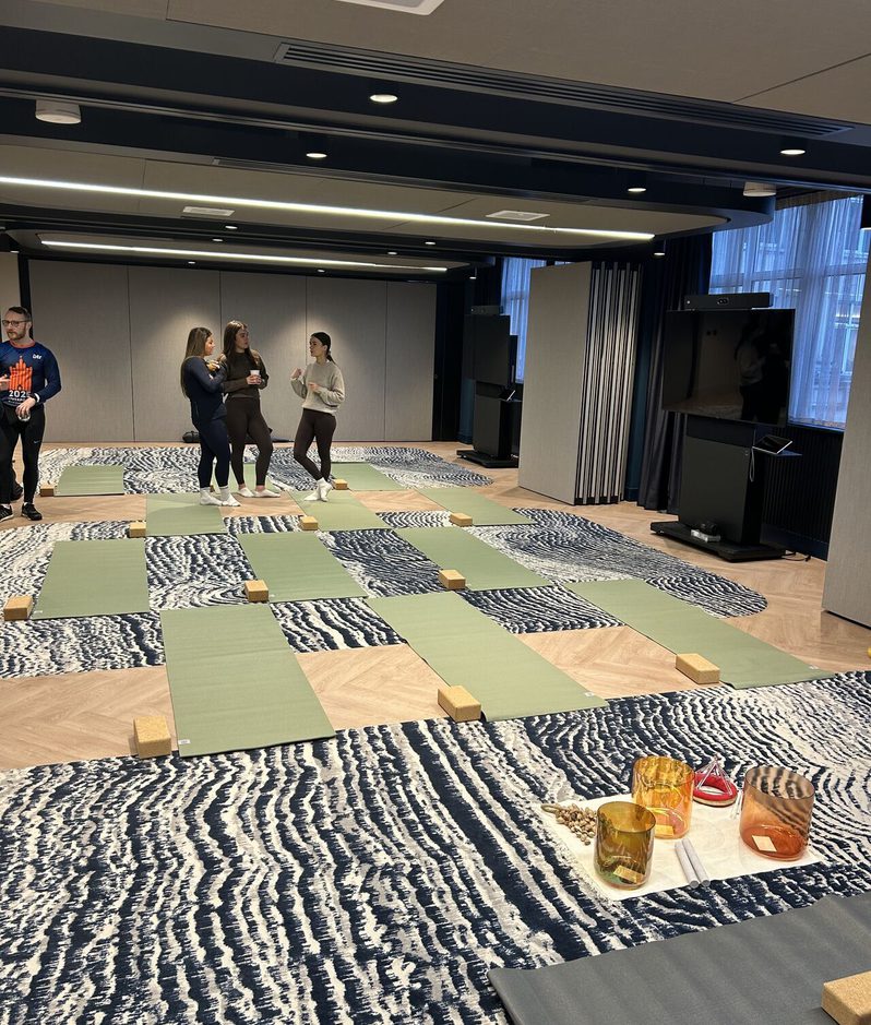 Several people stand and chat in a modern room with green yoga mats arranged in a grid, cork blocks, and a blue patterned carpet; a large screen and equipment against the wall.