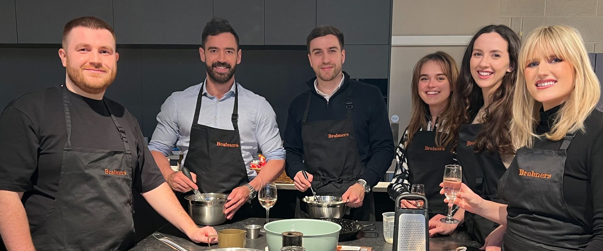 Six people standing around a kitchen counter, smiling, with cooking utensils, bowls, and glasses of wine in a modern kitchen.