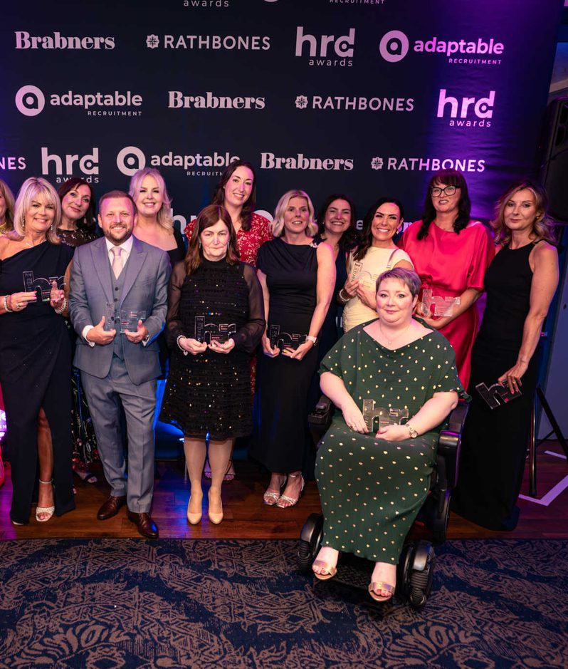 Group of award winners posing with glass trophies in front of a blue backdrop branded with hrd awards, adaptable recruitment, Brabners and Rathbones logos; a person in a green polka-dot dress sits in a wheelchair.