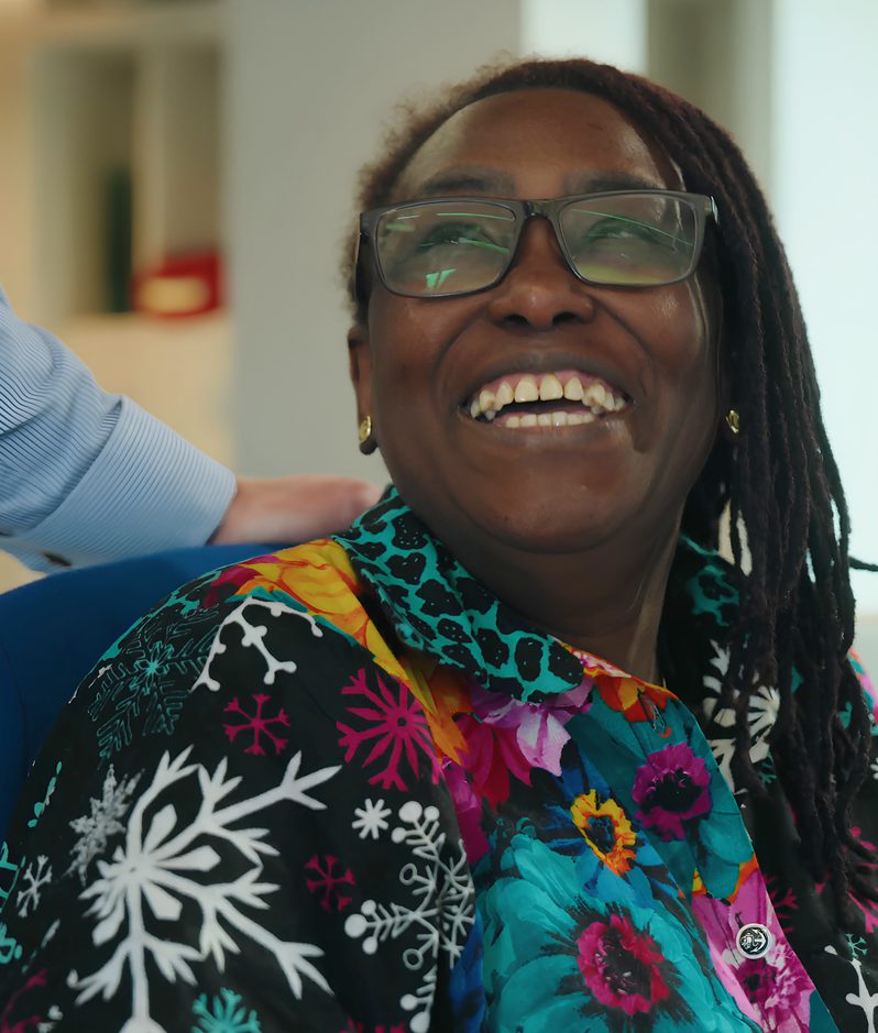 Joyful person with glasses and dreadlocks, wearing a colourful floral-patterned shirt, smiling broadly in an office while holding a lined notebook.