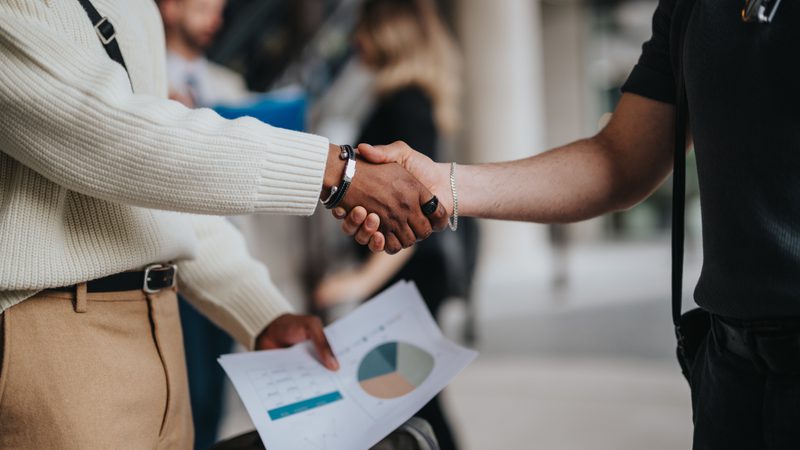 Two people shake hands, one holding a sheet of paper with charts, in a professional setting with blurred individuals in the background.