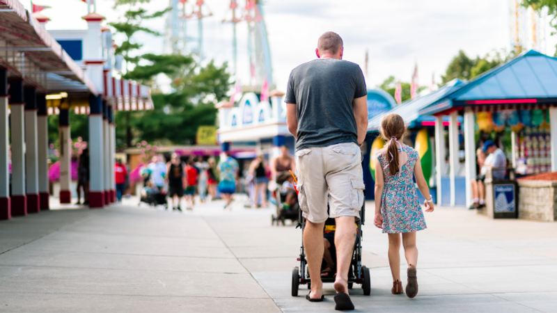 A person pushing a stroller walks with a young girl down a sunny amusement park walkway lined with colourful stalls and rides in the background.