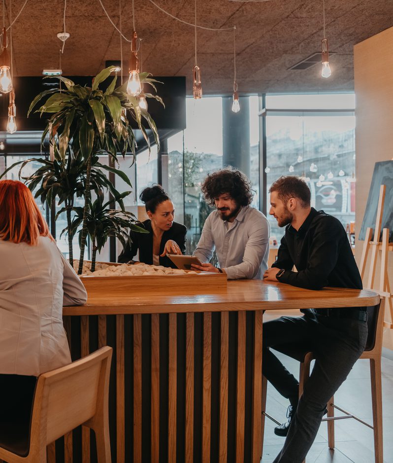 Six people sitting and standing around a wooden table, engaged in conversation, with large windows, a tall plant, and warm lighting in a modern office space.