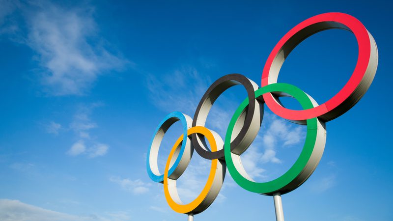 The Olympic rings sculpture with five interlinked rings in blue, yellow, black, green, and red against a blue sky with scattered clouds.