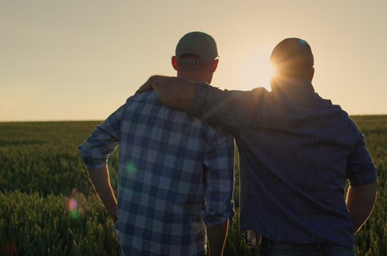 A senior farmer family business owner puts his arm around his son and faces the sunset across fields