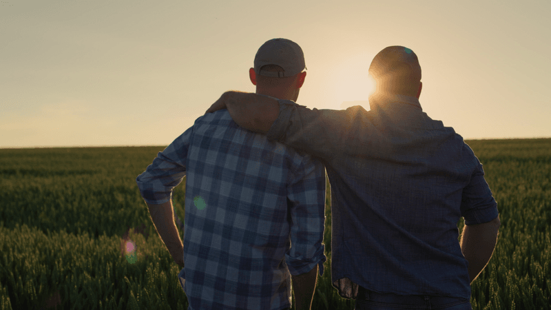 A senior farmer family business owner puts his arm around his son and faces the sunset across fields