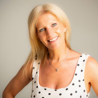 Smiling blonde person with blue eyes wearing a white sleeveless polka dot top, silver necklace, posing against a plain light background