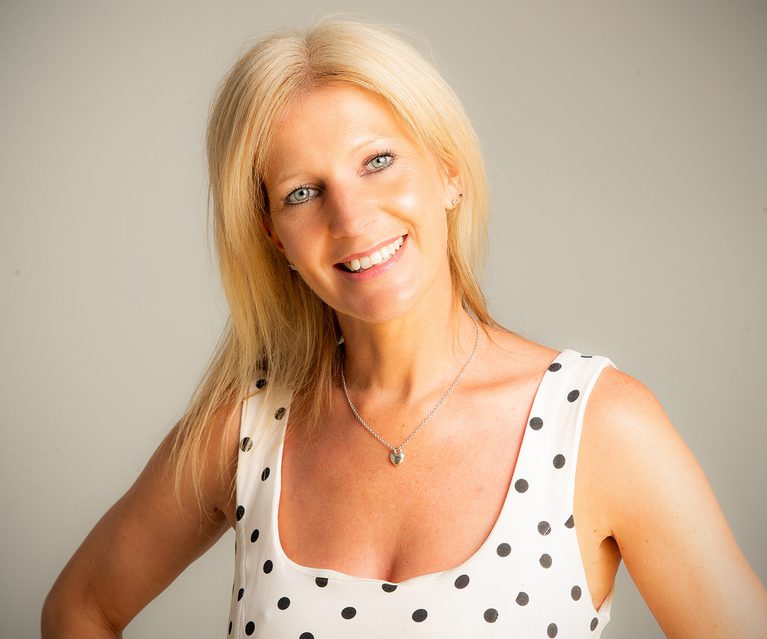 Smiling blonde person with blue eyes wearing a white sleeveless polka dot top, silver necklace, posing against a plain light background