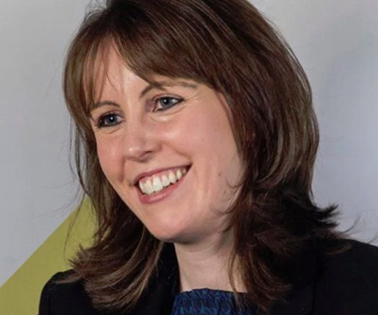 A smiling woman with shoulder-length brown hair, wearing a black blazer and a blue top, posed against a light grey background.