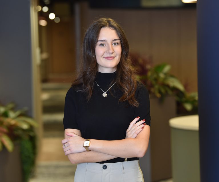 A young woman with long brown hair, wearing a black top and beige trousers, stands confidently with arms crossed in an indoor setting with plants behind her.