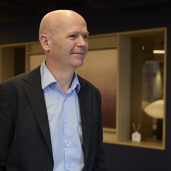 Bald person in a light blue shirt and dark blazer stands indoors, smiling softly, with a modern interior featuring warm lighting and wooden panels behind.