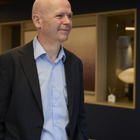 Bald person in a light blue shirt and dark blazer stands indoors, smiling softly, with a modern interior featuring warm lighting and wooden panels behind.